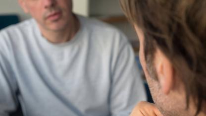 Zwei m&auml;nnlich gelesene Personen sitzen sich in einem ernsten Gespr&auml;ch gegen&uuml;ber.